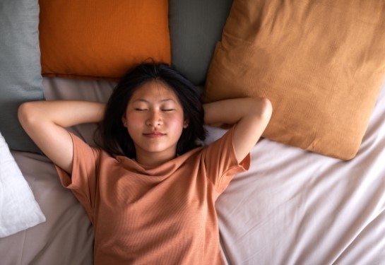 Person lying on a bed with hands behind head, eyes closed, resting among neutral-toned pillows. 