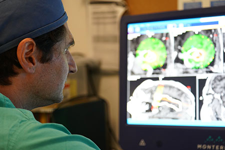 A health care provider in scrubs and a cap examines patient brain scans on monitors. 