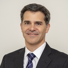 Professional headshot of a person wearing a dark suit, white dress shirt and striped tie against a light gray background. The person is facing the camera.