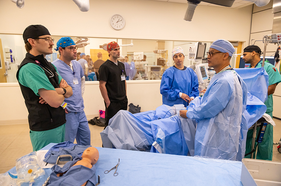 Faculty and students gather around a patient bed in an operating room simulation during a surgical training session.