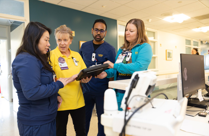 Four people stand at a nurses station and look at a tablet computer.