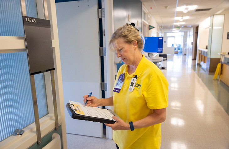 Volunteer writes on a clipboard while standing outside a patient room in a hospital hallway