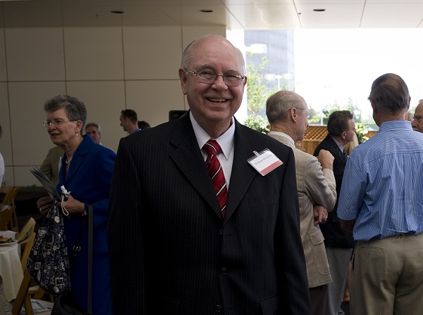 William McGowan stands at a UC Davis Health event wearing a dark suit and red striped tie while attendees gather in the background.