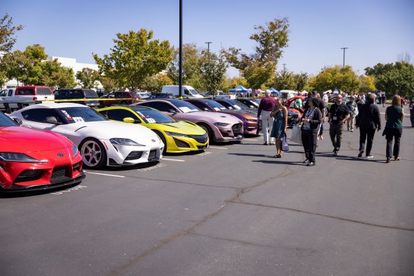 Sports cars lined up at a car show as attendees walk through the display on a sunny day. 