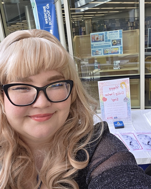 A person dressed in a Dolly Parton-inspired outfit stands behind a table featuring signs, QR codes and information about Dolly Parton's Imagination Library outside the Blaisdell Medical Library.