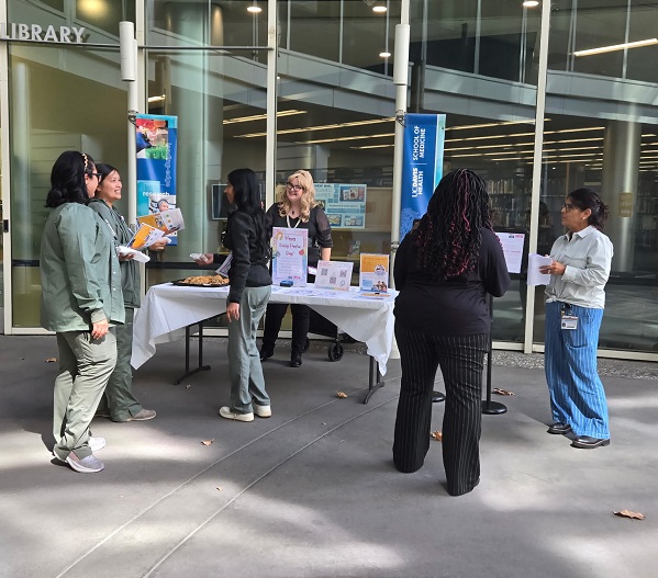 Several employees stand and talk around a table outside the Blaisdell Medical Library during a Dolly Parton Day pop-up event. The table includes informational displays and giveaway items.