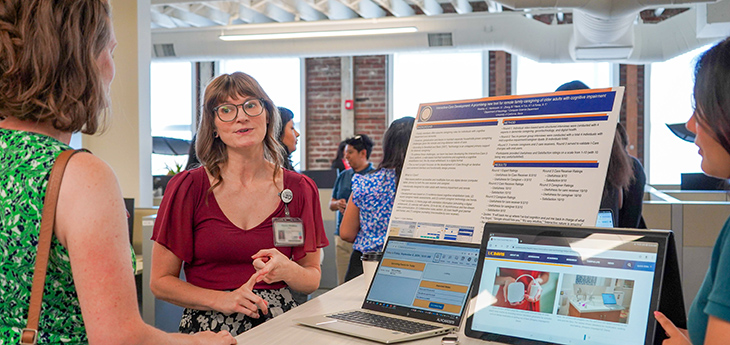 Professor Alyssa Weakley standing next to a poster and speaking to someone in the audience.