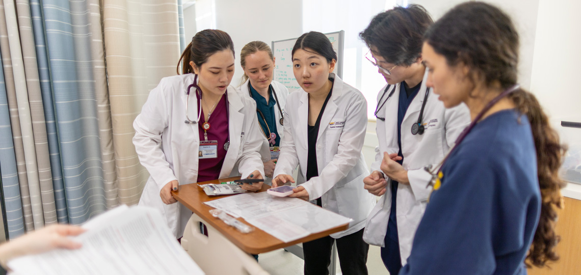 Health professions students in scrubs and white coats work together around a patient bed during a clinical simulation exercise.