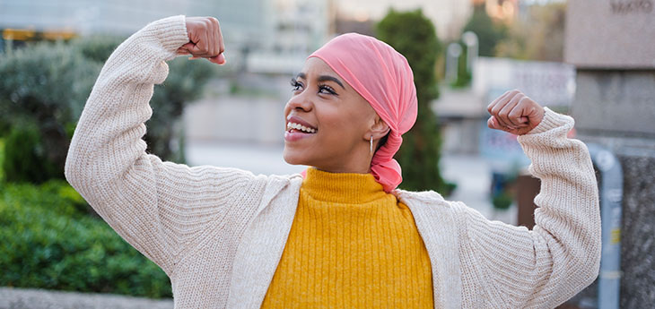A woman wearing a pink head scarf stands in an urban area with her arms raised in triumph. 