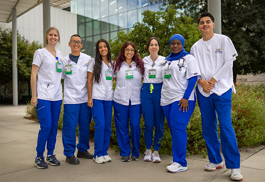 Seven nursing students in white scrub tops and blue pants stand together outdoors in front of a modern campus building.