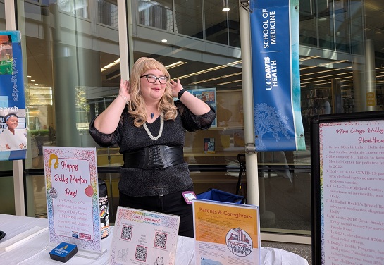 A person wearing a blonde wig and glasses takes a selfie in front of a Dolly Parton Day display table outside the Blaisdell Medical Library. 