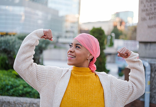 A woman wearing a pink head scarf stands in an urban area with her arms raised in triumph. 