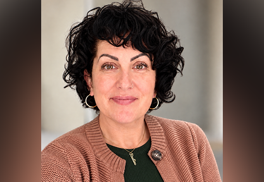 Professional headshot of a person with short curly dark hair, wearing hoop earrings, a green top and a light brown cardigan.