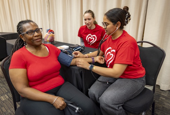 A participant sits in a chair while a volunteer wearing a red shirt uses a blood pressure cuff.