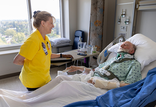 Volunteer stands beside a hospital bed and talks with a patient in a hospital room