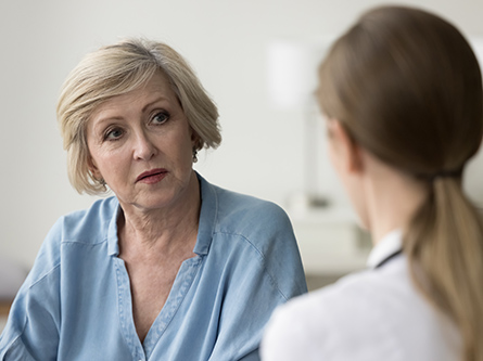 A woman in a blue shirt speaks with a physician in a white coat in a medial office.
