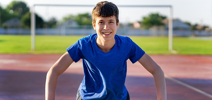 Boy with blue shirt standing on track leaning on rail with goal posts and grass behind him.