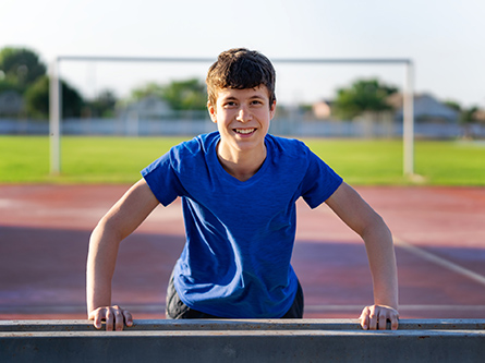 Boy with blue shirt standing on track leaning on rail with goal posts and grass behind him.