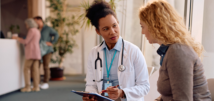 A female doctor wearing a white coat with a stethoscope is sitting next to a woman with long curly hair in a medical office explaining something.