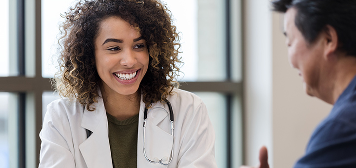 A female doctor in a white coat sits at a table, smiling and speaking with a man in a blue shirt.