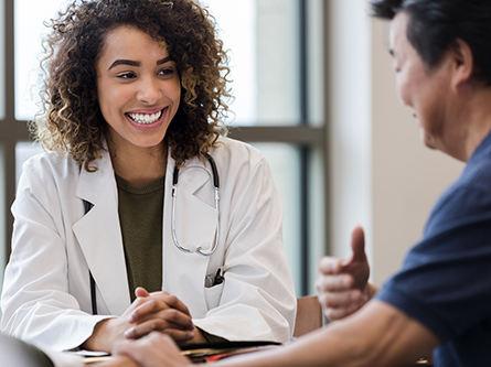 A female doctor in a white coat sits at a table, smiling and speaking with a man in a blue shirt.