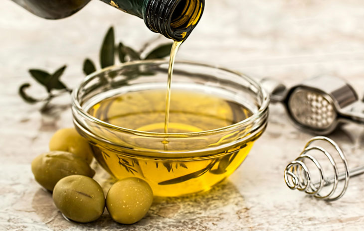 bottle of olive oil being poured into a clear bowl with a couple green olives next to it