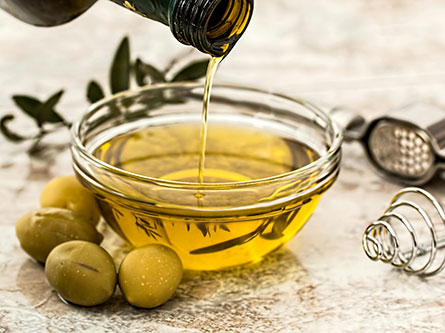 bottle of olive oil being poured into a clear bowl with a couple green olives next to it