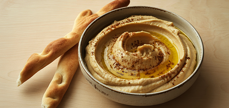 A bowl of swirled hummus with olive oil and ground paprika beside two bread sticks