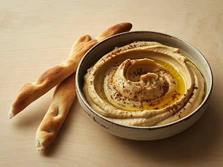 A bowl of swirled hummus with olive oil and ground paprika beside two bread sticks