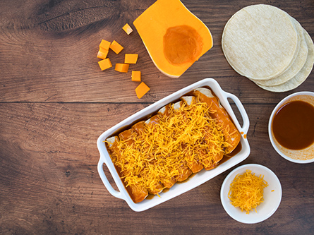 White baking dish with butternut squash enchiladas topped with cheese, surrounded by tortillas, squash cubes and sauce on a wooden table.