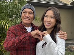 Hai and daughter Viana stand close together outdoors in front of a house and leafy plants. They are forming a heart shape with their hands at the center of the image.