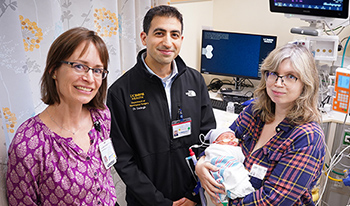 Female and male doctors in casual clothes stand next to a mother holding her infant baby in a hospital room.