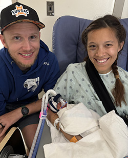 Two adults sit closely together in a hospital room. One adult is holding a newborn wrapped in blankets and connected to medical tubes.