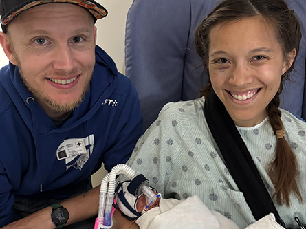 Two adults sit closely together in a hospital room. One adult is holding a newborn wrapped in blankets and connected to medical tubes.