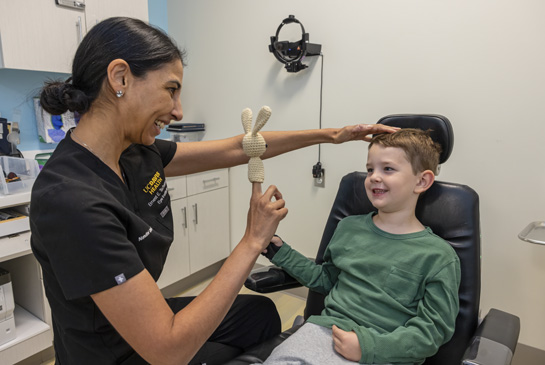 A female eye doctor at UC Davis Health holds up a small knitted bunny finger puppet to engage a smiling young boy sitting in an exam chair.