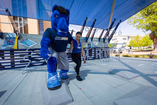 The UC Davis Health mascot and a woman doing lunges at the fitness court.