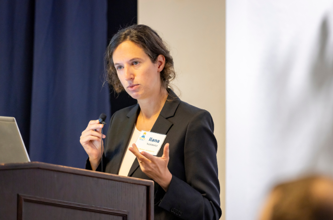 A person in a dark suit speaks at a podium while holding a microphone and gesturing with a hand. A name tag with 'Ilana' is visible on their jacket.