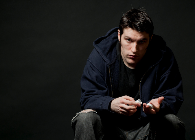 A white man with dark hair and black sweatshirts sits while pouring out a pill bottle.