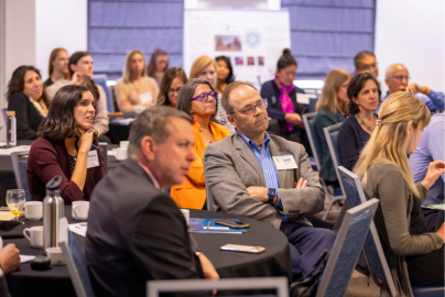 A diverse group of people seated at tables, attentively listening to a presentation. Some individuals have name tags.