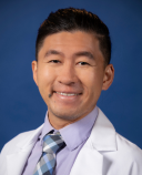 Smiling person in a white lab coat with a light blue dress shirt and striped tie, posed against a solid blue background.