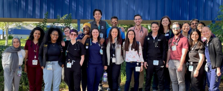 Outdoors under trees and next to a school, 20 UC Davis family medicine residents pose for a group photo.
