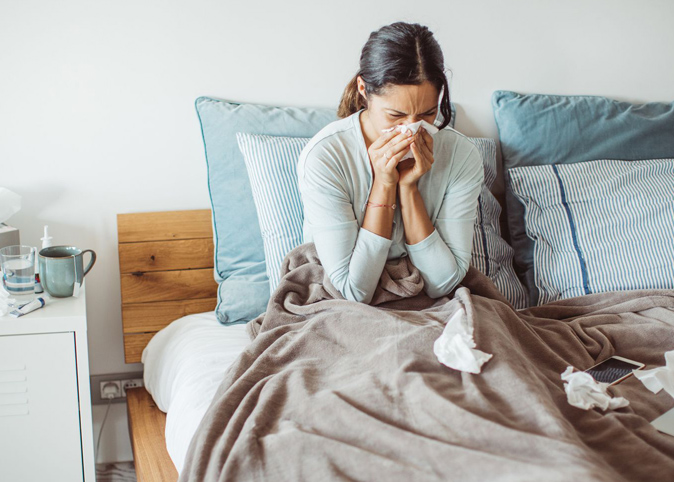 A woman in bed blowing her nose next to a nightstand with medicine, tissues, and tea on it.