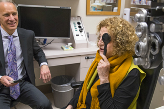 A patient covers one eye during a vision exam while a provider looks on in an eye care clinic.