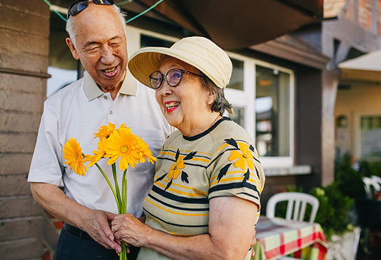 Elderly couple standing on a porch, holding flowers.