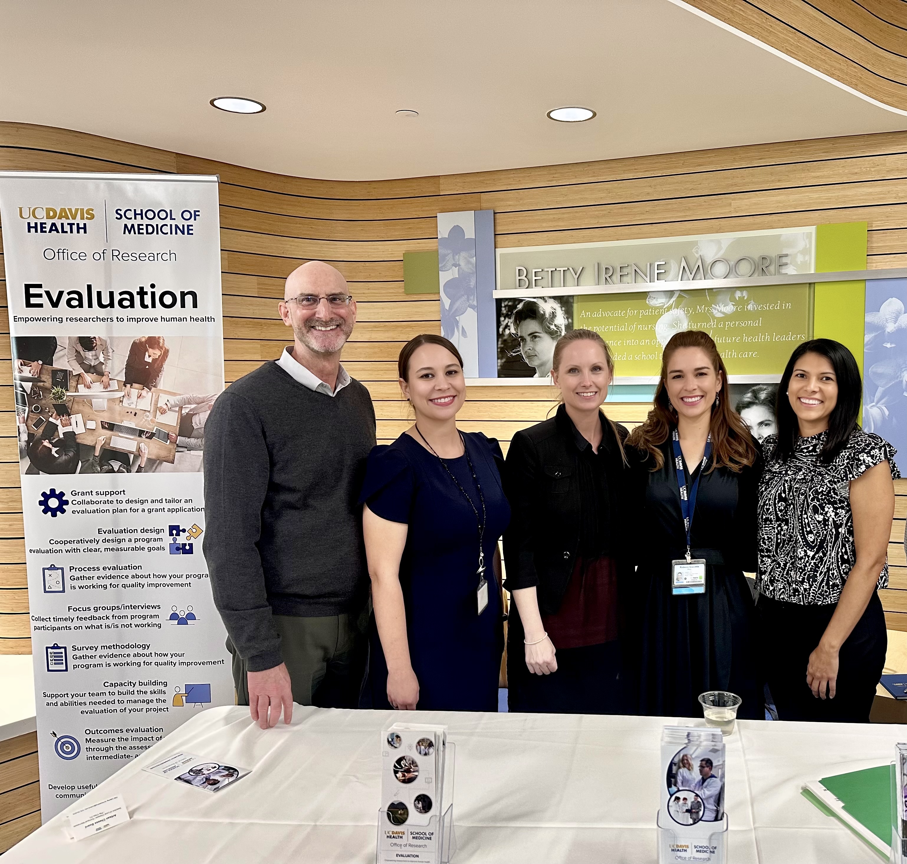 four professionals stand behind a table, smiling at a camera, next to a tall banner that reads "Evaluation"