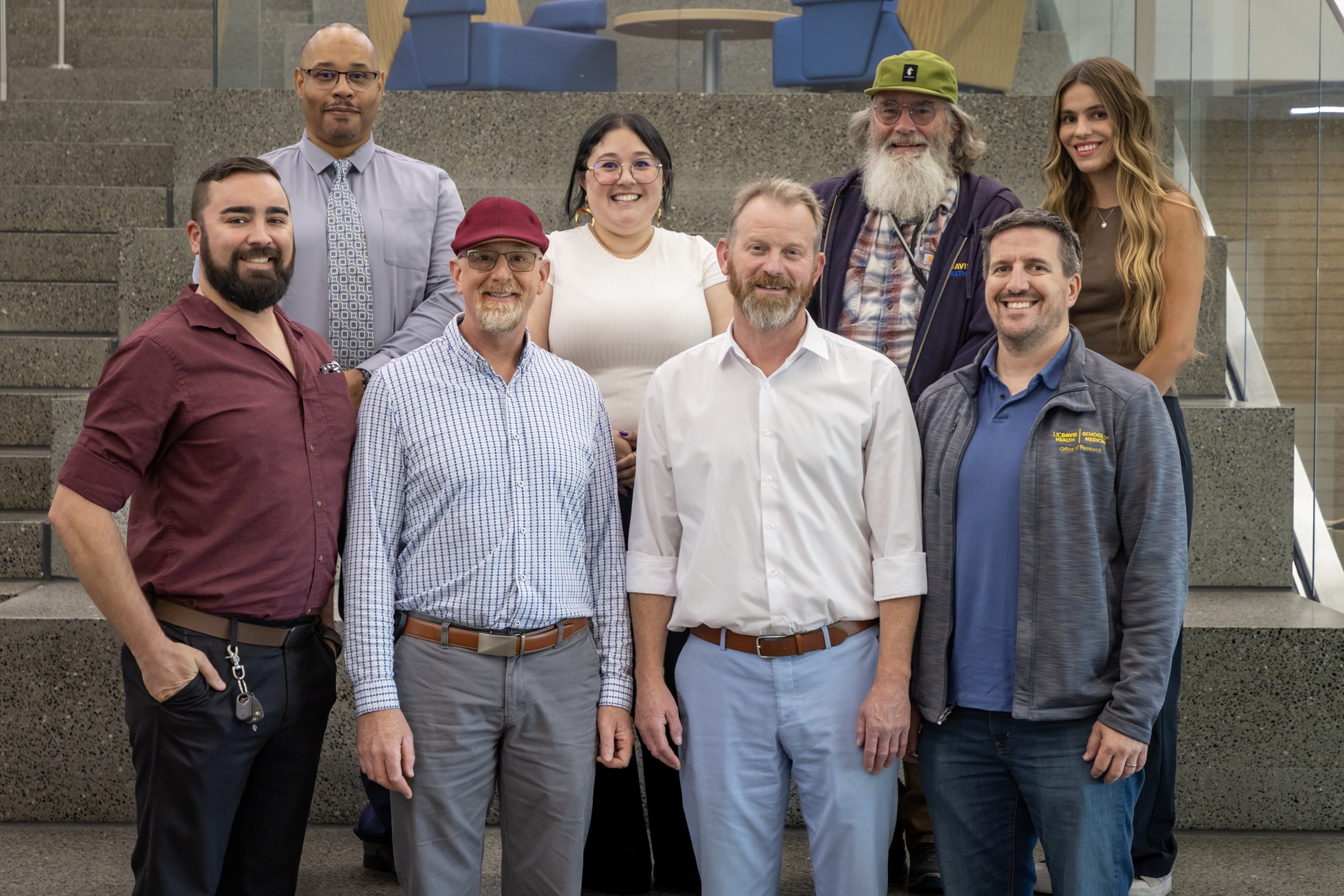 Group photo of eight staff members standing on indoor concrete steps in a modern academic building. 