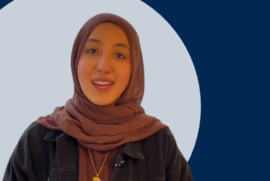 A woman wearing a brown hijab and dark jacket speaks to the camera against a light gray circular background with a navy blue backdrop.