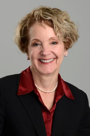 Studio portrait of Allison Brashear wearing a black blazer over a red blouse with a pearl necklace, photographed against a neutral gray background.