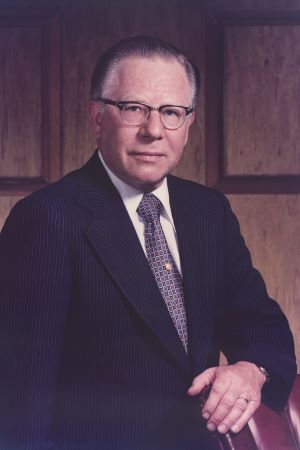 Formal painted portrait of C. John Tupper wearing a dark suit and glasses, seated indoors against a wood-paneled background.