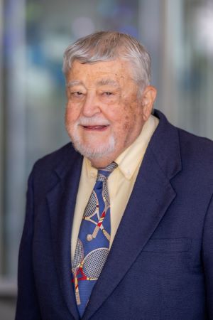 Portrait of Carroll Cross wearing a blue jacket, light-colored shirt, and patterned tie, standing indoors with a softly blurred background.